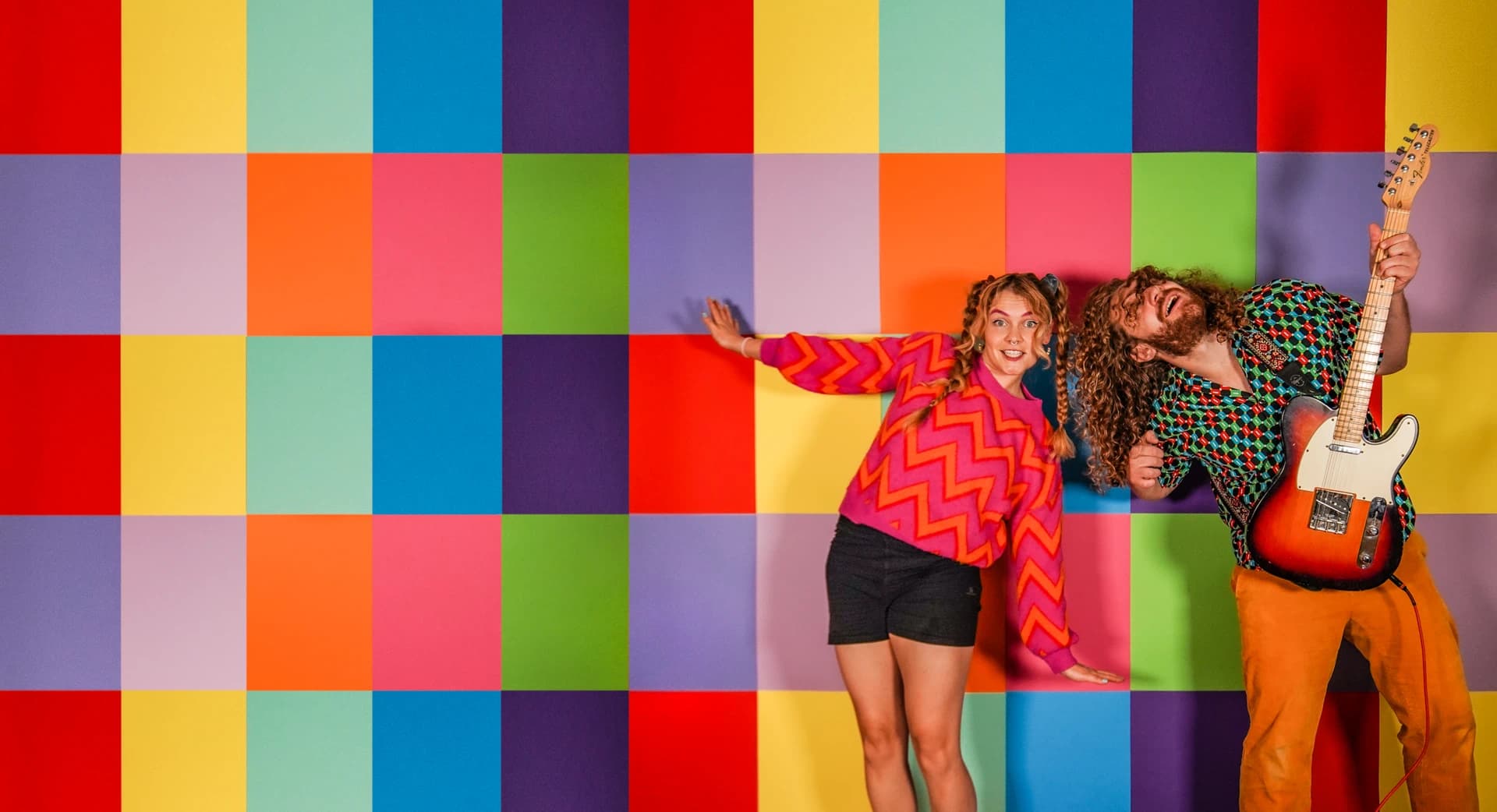 Promotional image of UK / Germany pop duo, Fruit Sharks, standing in front of a colourful background, posing and playing instruments